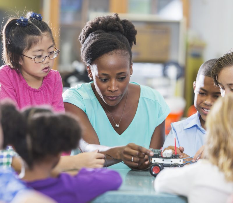 An African American teacher with a group of elementary school children in a science classroom working on a robotics project. The woman is helping a boy attach wires to a model vehicle while an Asian girl watches.